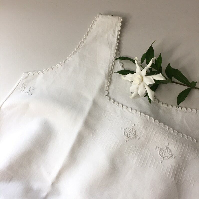 May include: White linen sleeveless top with scalloped edges and embroidered details. The top has the initials "B & L" embroidered on the front.