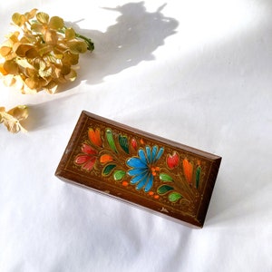 May include: A wooden box with a floral design painted on the lid. The flowers are red, blue, green, and yellow. The box is closed.