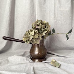 May include: A copper-toned, vintage-style vase with a long wooden handle, filled with dried hydrangeas. The vase sits on a white cloth, with a small sprig of the same flowers nearby. The overall aesthetic is rustic and evokes a sense of antique charm.