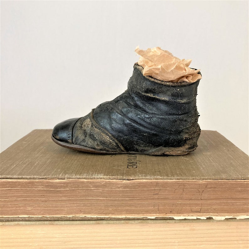 May include: A black leather baby shoe with a worn appearance. The shoe is sitting on top of a stack of books.