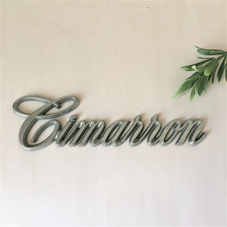 May include: Silver metal car emblem with the word "Cimarron" in a cursive script font.