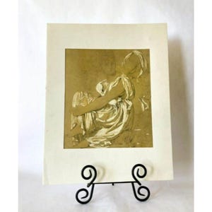 Puede incluir: Una impresión en tonos sepia de una figura sentada con un drapeado fluido, exhibida en un caballete de hierro forjado negro. La obra de arte está montada sobre papel color crema, creando una estética clásica. Los detalles de la impresión son visibles.
