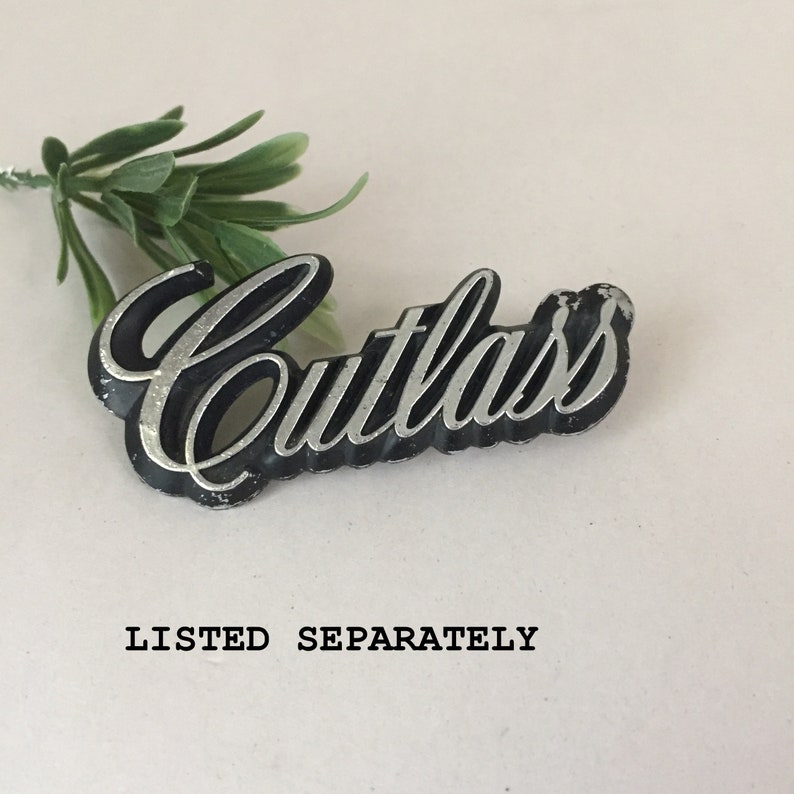 May include: A black and silver car emblem with the word "Cutlass" in a cursive font.
