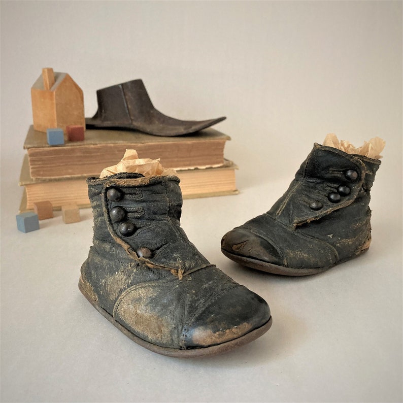 May include: A pair of black leather baby boots with button closures. The boots are worn and have a distressed look. The boots are sitting on a stack of books.