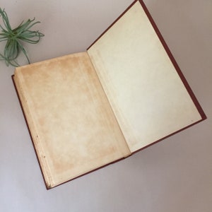 May include: An open book with blank pages and a red cover. The book is lying flat on a light brown surface.