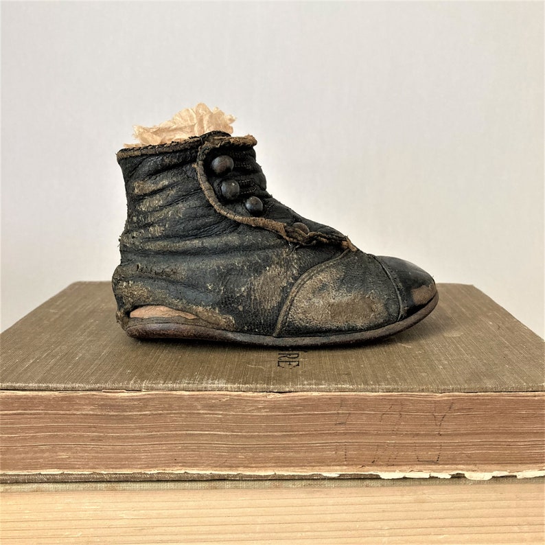 May include: A vintage black leather baby boot with button closures. The boot is sitting on top of a stack of old books.
