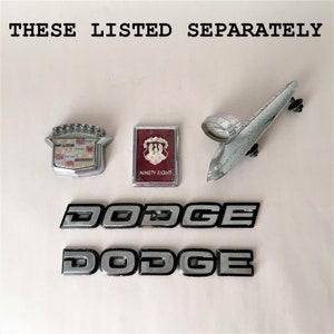 May include: A collection of vintage car emblems, including two silver Dodge emblems, a silver Cadillac emblem, a silver and red Ninety Eight emblem, and a silver car hood ornament.