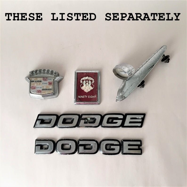 May include: A collection of vintage car emblems, including two silver Dodge emblems, a silver Cadillac emblem, a silver and red Ninety Eight emblem, and a silver car hood ornament.