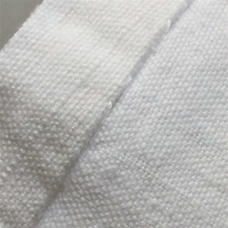 May include: Close-up of a white fabric with a textured weave. The fabric is folded over itself, showing the seam.