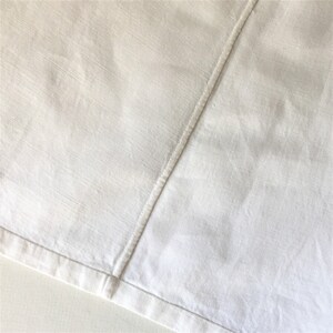 May include: A close-up of a white fabric with a seam running diagonally across the surface. The fabric has a subtle texture and a slightly wrinkled appearance.