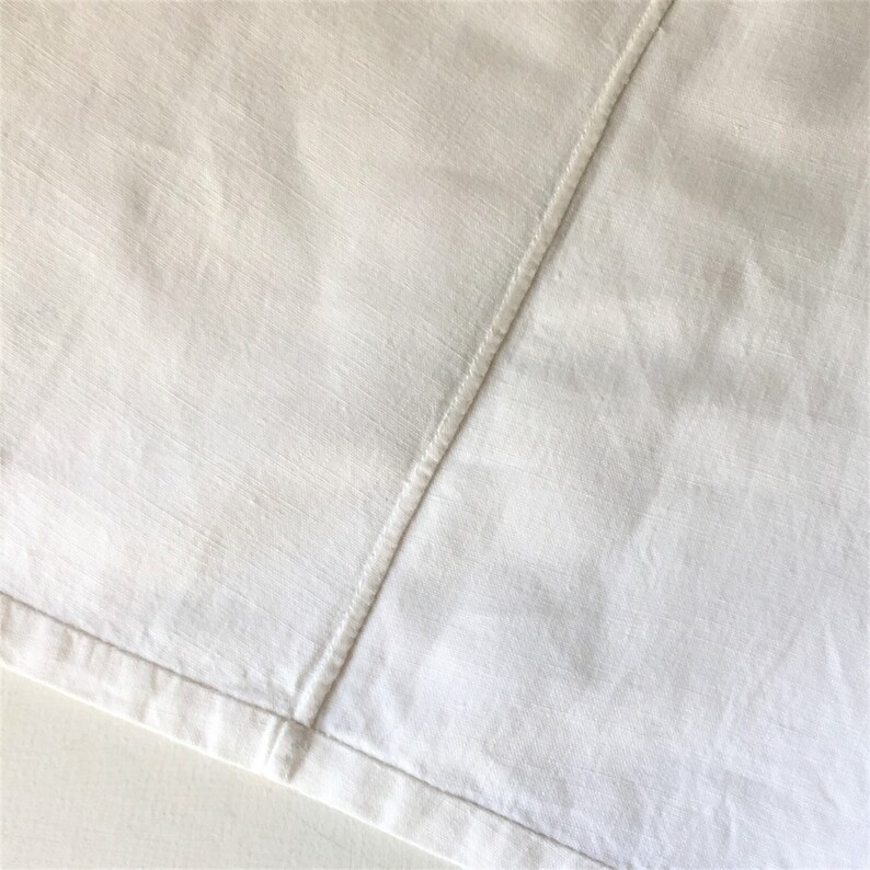 May include: A close-up of a white fabric with a seam running diagonally across the surface. The fabric has a subtle texture and a slightly wrinkled appearance.
