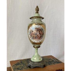 May include: A vintage, light green porcelain urn with a gold-trimmed oval scene depicting figures in a garden. The urn has a matching lid and sits on a pedestal base. The base is a dark metal color.