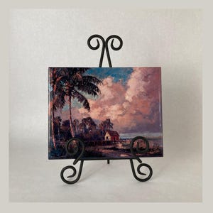 May include: A ceramic tile with a painting of a tropical scene. The painting depicts a palm tree in the foreground, with a small house and a cloudy sky in the background. The tile is on a black metal easel.
