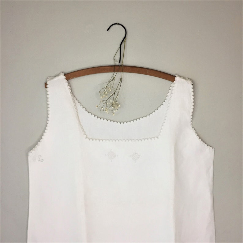 May include: White sleeveless top with a scalloped neckline and embroidered details. The top is hanging on a wooden hanger.