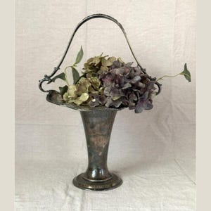 May include: A vintage silver-toned vase with a handle, filled with dried hydrangeas in shades of green and purple. The vase has a flared top and a pedestal base, perfect for floral arrangements. The handle is arched and decorative.