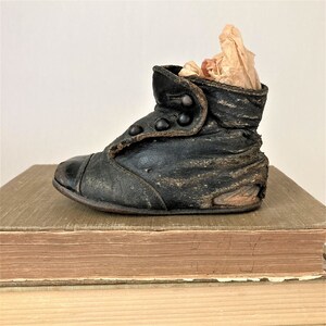 May include: A vintage black leather baby shoe with button closures. The shoe is worn and has a distressed look. It is sitting on a stack of old books.