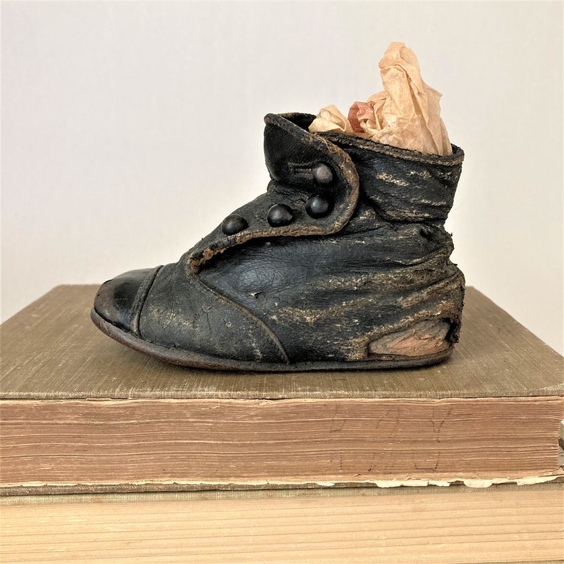 May include: A vintage black leather baby shoe with button closures. The shoe is worn and has a distressed look. It is sitting on a stack of old books.