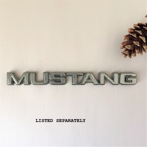 May include: Silver metal car emblem with the word "MUSTANG" in raised letters.