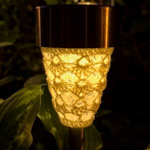 Puede incluir: Una lámpara solar de jardín con una parte superior metálica cilíndrica y un cuerpo de vidrio cónico envuelto en una funda de ganchillo de color crema con patrón abierto. La luz emite un brillo cálido a través del diseño texturizado.