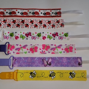 May include: Six baby pacifier clips with various designs. The clips feature red, white, purple, and yellow ribbons with ladybugs, butterflies, dragonflies, and bees. Each clip has a plastic clasp and a white elastic loop for attaching to a pacifier.