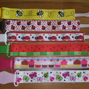 May include: Six colorful pacifier clips with different patterns, including bees, ladybugs, watermelons, cupcakes, and butterflies. Each clip has a white elastic strap.