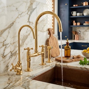 Può includere: Un set di rubinetti da cucina in oro lucido con bocca ad arco alto, un rubinetto secondario più piccolo e comandi a croce. Il rubinetto è montato su un piano di lavoro in marmo bianco con un lavello in rame, con acqua che scorre.