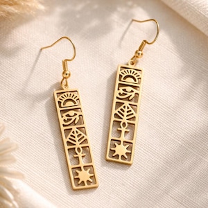 Könnte beinhalten: Ein Paar goldfarbene Ohrhänger mit rechteckigen Anhängern. Jeder Anhänger zeigt eine vertikale Reihe ausgeschnittener Symbole, darunter eine aufgehende Sonne, das Auge des Horus, ein Blatt, ein Ankh und ein Stern, befestigt an einfachen Ohrhaken.