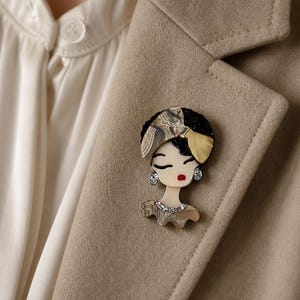 Peut inclure: Une broche en forme de visage stylisé aux yeux fermés et aux lèvres rouges. La figure porte un foulard texturé dans des tons beige et noir, des accents dorés et de petites boucles d'oreilles circulaires. Un collier argenté orne le cou.
