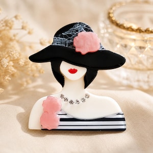 Peut inclure: Une broche représentant une silhouette portant un chapeau noir à larges bords avec un motif en dentelle blanche et une fleur rose. La silhouette a un teint blanc, des lèvres rouges, un collier en strass et un haut rayé noir et blanc avec des détails floraux roses.