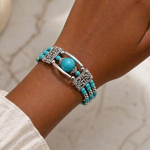 Op de afbeelding: Een armband met meerdere strengen, voorzien van turquoisekleurige ronde kralen en kleine zilverkleurige metalen kralen. Het ontwerp bevat rechthoekige zilverkleurige filigraan tussenstukken en een centrale, grotere turquoisekleurige bol met zilverkleurige accenten.