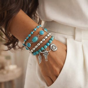 Op de afbeelding: Een set van vier gelaagde armbanden met turkooiskleurige kralen, zilverkleurige metalen bollen en ovale turkooiskleurige stenen. De armbanden bevatten zilverkleurige bedels in de vorm van een veer, een stierenschedel en een cowboyhoed.