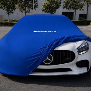 以下が含まれることがあります： 鮮やかな青いカーカバーで部分的に覆われた白いスポーツカー。青い生地のボンネット部分には白いAMGのロゴがプリントされています。車両のフロントグリルが見えており、メルセデス・ベンツのエンブレムとAMGのバッジが確認できます。