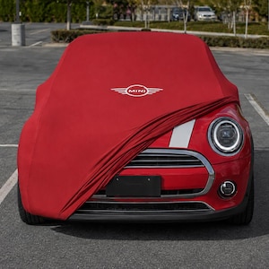 May include: A red car cover with the white MINI logo printed on the front, partially draped over the hood and windshield of a red car. The cover features a smooth fabric texture and is positioned to protect the front section of the vehicle.