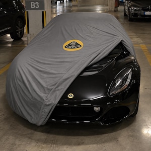 May include: A black Lotus sports car partially covered by a grey fabric car cover. The cover features a yellow and green Lotus logo on the bonnet, while the car's front bumper displays a smaller matching Lotus emblem and a visible headlight.