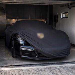 以下が含まれることがあります： ガレージ内に駐車された黒いスポーツカーで、黒い布製の専用カーカバーがかけられています。カバーの下からは、フロントヘッドライトと光沢のある黒いバンパーの一部が見えており、ボンネット部分にはぼやけたエンブレムがあしらわれています。