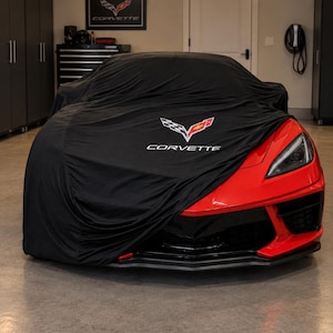 May include: A bright red sports car partially covered by a black fabric car cover. The cover features the white and red Corvette logo and the text CORVETTE printed in white. The car has a sharp front bumper design and a visible angular headlight.