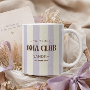 Op de afbeelding: Een witte keramische mok met verticale lichtpaarse strepen, voorzien van de donkerbruine tekst NEW MEMBER OMA CLUB, de naam SANDRA en est. März 2027. Een bijpassende kaart met de tekst Willkommen im besten Club der Welt! staat achter de mok.