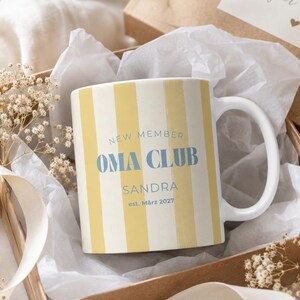 Puede incluir: Una taza de cerámica blanca con rayas verticales amarillas, con el texto azul NEW MEMBER OMA CLUB, el nombre SANDRA y la fecha est. März 2027. La taza está colocada en una caja de regalo con papel de seda blanco y flores secas.