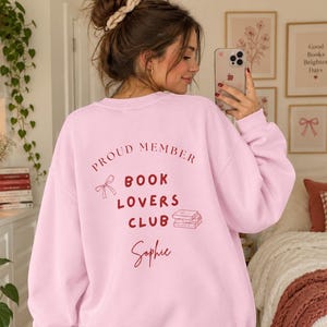 Op de afbeelding: Een lichtroze sweater met ronde hals en donkerrode tekst op de achterkant met de tekst PROUD MEMBER BOOK LOVERS CLUB, voorzien van een kleine lijntekening van een strik en een stapel boeken, gevolgd door de naam Sophie in cursief schrift.