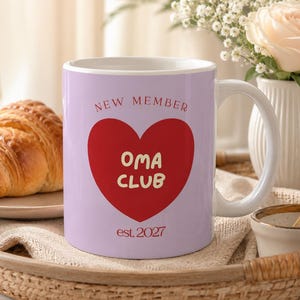 May include: A light purple ceramic mug featuring a large red heart graphic on the front. Inside the heart, the text reads "Oma CLUB" in yellow, with "NEW MEMBER" arched above and "est. 2027" printed in red below the heart shape.