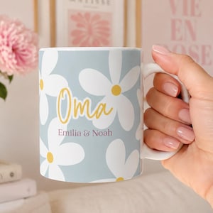 Puede incluir: Una taza de cerámica con fondo azul claro que presenta un patrón de margaritas blancas con centros amarillos. El texto "Oma" está impreso en una fuente cursiva amarilla, seguido de los nombres "Emilia & Noah" en una fuente sans-serif morada oscura.