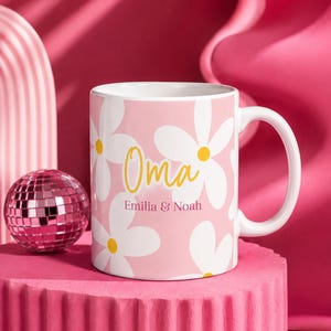Puede incluir: Una taza de cerámica blanca con fondo rosa que presenta un patrón repetitivo de margaritas blancas con centros amarillos. El texto "Oma" está impreso en una fuente cursiva amarilla sobre el texto "Emilia & Noah" en una fuente serif rosa oscuro.