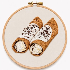 May include: A circular wooden embroidery hoop containing a fabric panel with a hand-stitched design of two cannoli. The pastries feature golden-brown shells, white cream filling, and dark brown chocolate chip accents rendered in textured thread.
