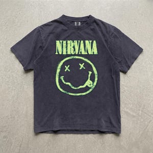 Peut inclure: Un t-shirt à manches courtes gris anthracite arborant le mot NIRVANA dans une police vert citron usée au-dessus d'un motif graphique assorti représentant un visage souriant avec des yeux en X et une bouche ondulée tirant la langue.
