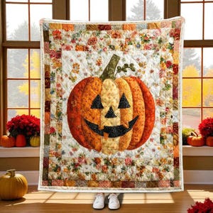 May include: A quilted throw blanket featuring a central jack-o'-lantern pumpkin graphic with a black triangular face and a wide grin. The pumpkin is surrounded by a white background with a floral pattern and a border of multicolored fabric squares.