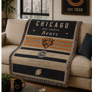 Puede incluir: Una manta tejida con el logotipo de los Chicago Bears, el nombre del equipo y texto que incluye NFC North, Est. 1920 y Soldier Field. El diseño incorpora rayas azul marino, naranja y crema con un gráfico central de cabeza de oso y un icono de balón.