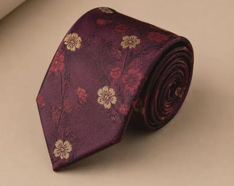 May include: A deep burgundy necktie featuring a woven pattern of small, five-petaled flowers in shades of gold and muted red. The fabric has a subtle sheen, and the tie is rolled into a coil, displaying the pointed tip and the floral motif.