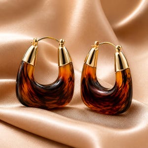 May include: A pair of U-shaped hoop earrings featuring a polished gold-tone metal top section and a curved, translucent brown tortoiseshell-patterned resin bottom section. The earrings have a hinged post closure mechanism at the top.