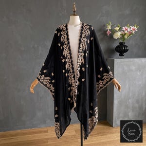 May include: A black open-front kimono featuring intricate beige floral embroidery along the edges and scattered throughout the fabric. The garment is displayed on a mannequin, showcasing a loose, flowing silhouette with wide sleeves and scalloped trim.