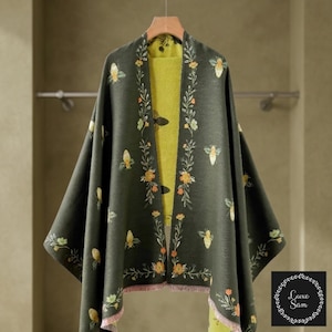 May include: A dark olive green open-front wrap featuring a repeating pattern of embroidered bees and floral vines. The garment has a contrasting bright lime green interior lining and a delicate light pink fringe along the bottom hem edges.